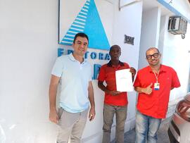 Retrato de Rômulo Vaz e Leandro Almeida com Neilson Santana, ex-pessoa em situação de rua, que exibe manuscrito de seu livro 'Vivências, memórias e escritas' em frente ao escritório da Editora Kelps